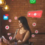 Młoda kobieta siedząca na krześle z telefonem w dłoniach. Wokół kobiety widoczne ikonki i emoji