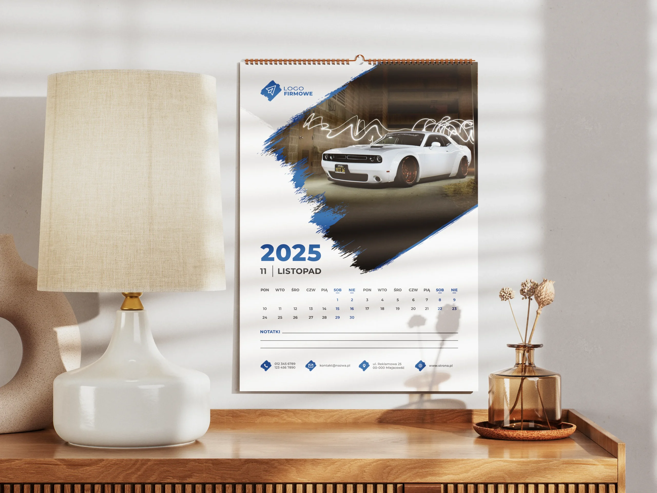 Kalendarz ścienny spiralowy wiszący na ścianie nad biurkiem z lampą i kwiatkiem